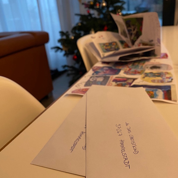 Briefumschläge, adressiert an das Christkind, dahinter liegen Wunschzettel auf dem Tisch und im Hintergrund steht der Weihnachtsbaum
